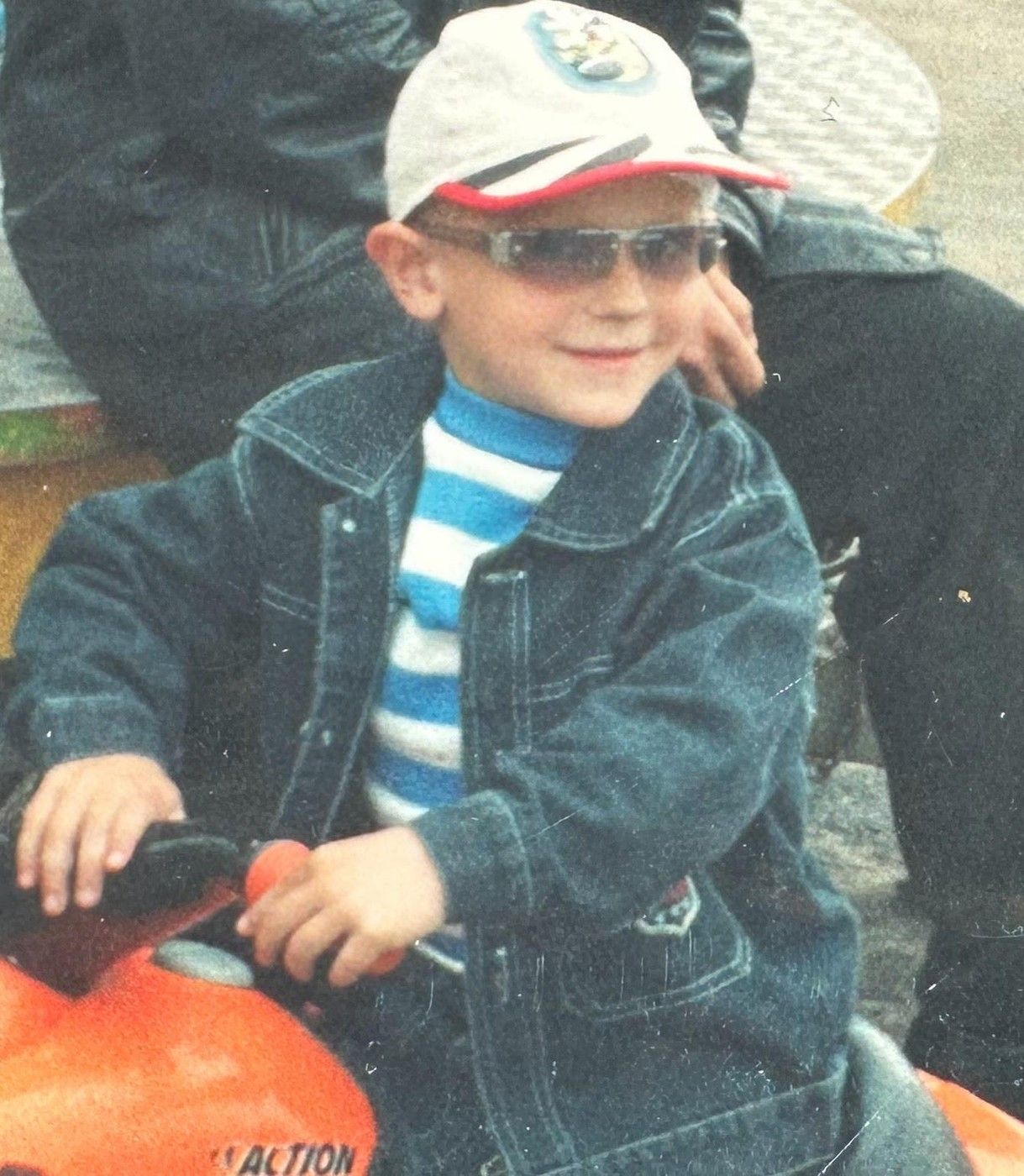 Taras de niño, con gorra, gafas de sol y chaqueta vaquera, sentado en una moto de juguete naranja